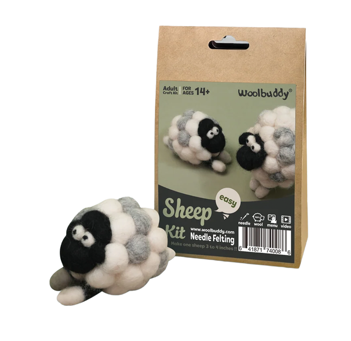 A Woolbuddy needle-felting sheep kit box stands behind a finished fluffy black, white, and gray sheep; the handmade craft display feels playful and inviting.