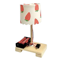  The kit set up includes a small tabletop lamp with a light wood base and stem, a white fabric shade printed with red strawberries, and a battery-powered circuit base holding red batteries and a black switch, isolated on a white background.