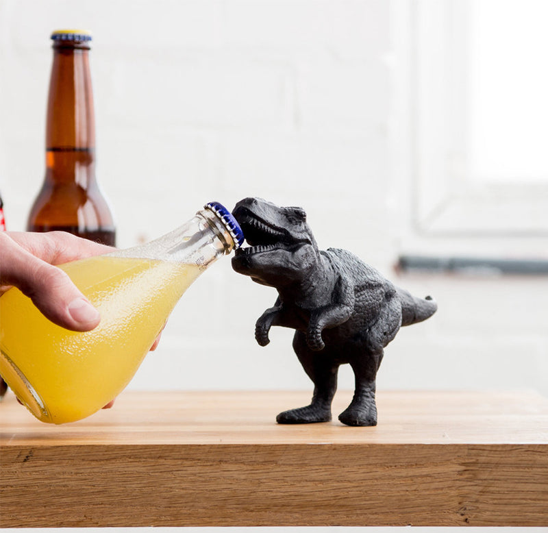 A hand holding a beverage in front of the dinosaur bottle opener positioned to open the bottle cap