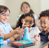 Three children in safety goggles watch blue foam bubble from a science kit on a classroom table, smiling with excitement as a teacher looks on.