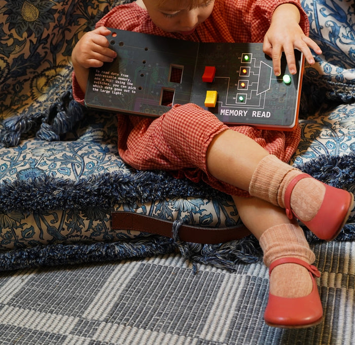 A child is sitting on a blanket with the Engineering Book For Big Babies opene showing two switches in red and yellow and four colored glowing lights.