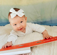 A baby wearing a white long-sleeve outfit and a white bow headband lies on a textured cream blanket, looking at a layout page from Engineering for Babies.