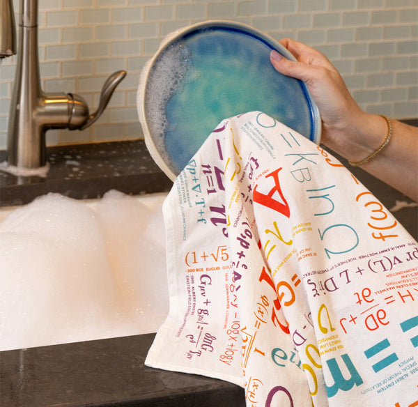 Person drying a dish with a colorful math-themed towel in a kitchen.