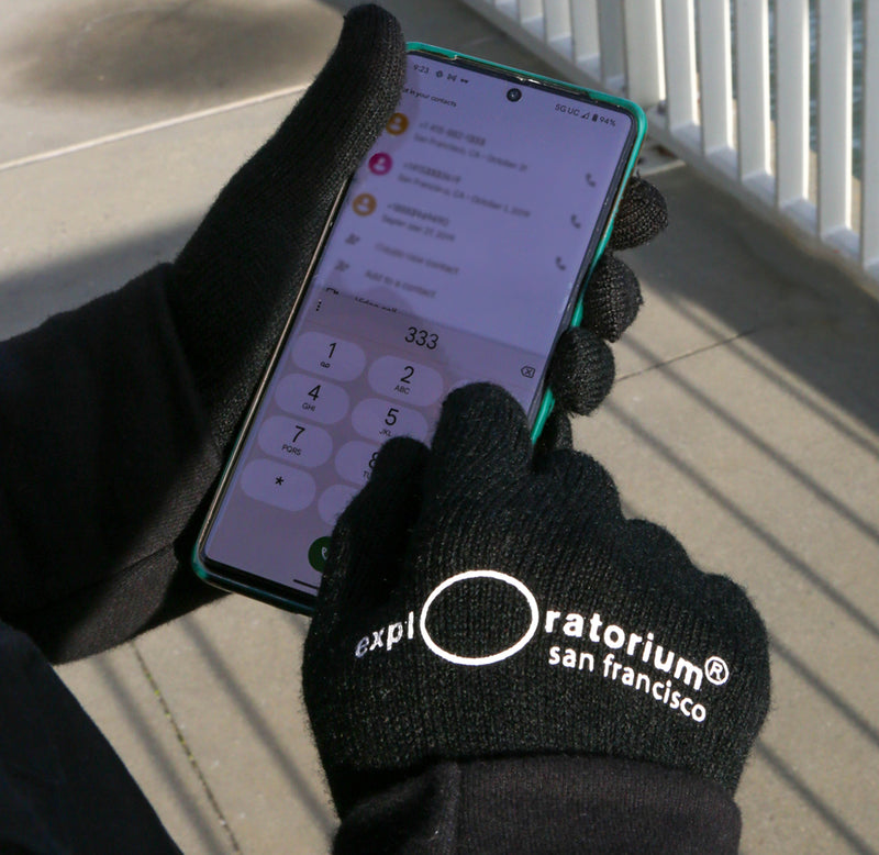 Image of black gloved hands with the exploratorium logo on the back of the gloves. Hands are holding mobile phone and typing a number on the screen. 