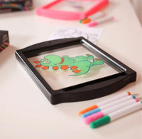 Coloring book with a green dragon drawing on a transparent sheet, surrounded by markers.
