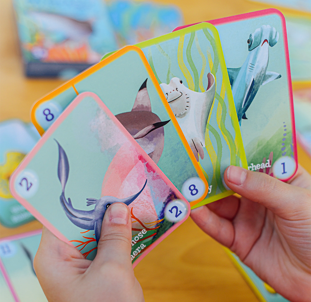 Hands fan out colorful ocean-themed game cards showing a whale, seal, and seabird, with numbered corners; playful educational activity on a table.