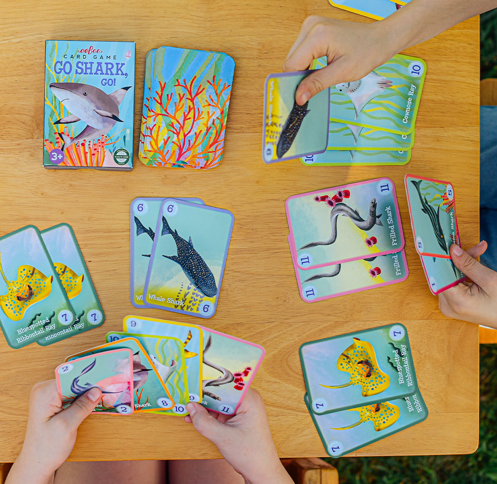 Hands arrange colorful ocean-animal cards on a wooden table, showing sharks, rays, and fish beside a “Go Shark Go!” game box; playful family game atmosphere.