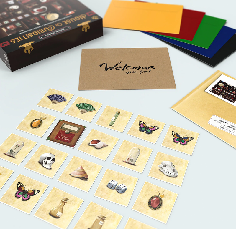 A styled flat-lay of a card game set on a light blue surface. The set includes a black box labeled 'House of Curiosities', a brown kraft card reading 'Welcome open first', multiple illustrated cards laid out in rows, colorful envelopes or card backs in yellow, red, green, blue, and black, and a gold-toned envelope with a printed insert.