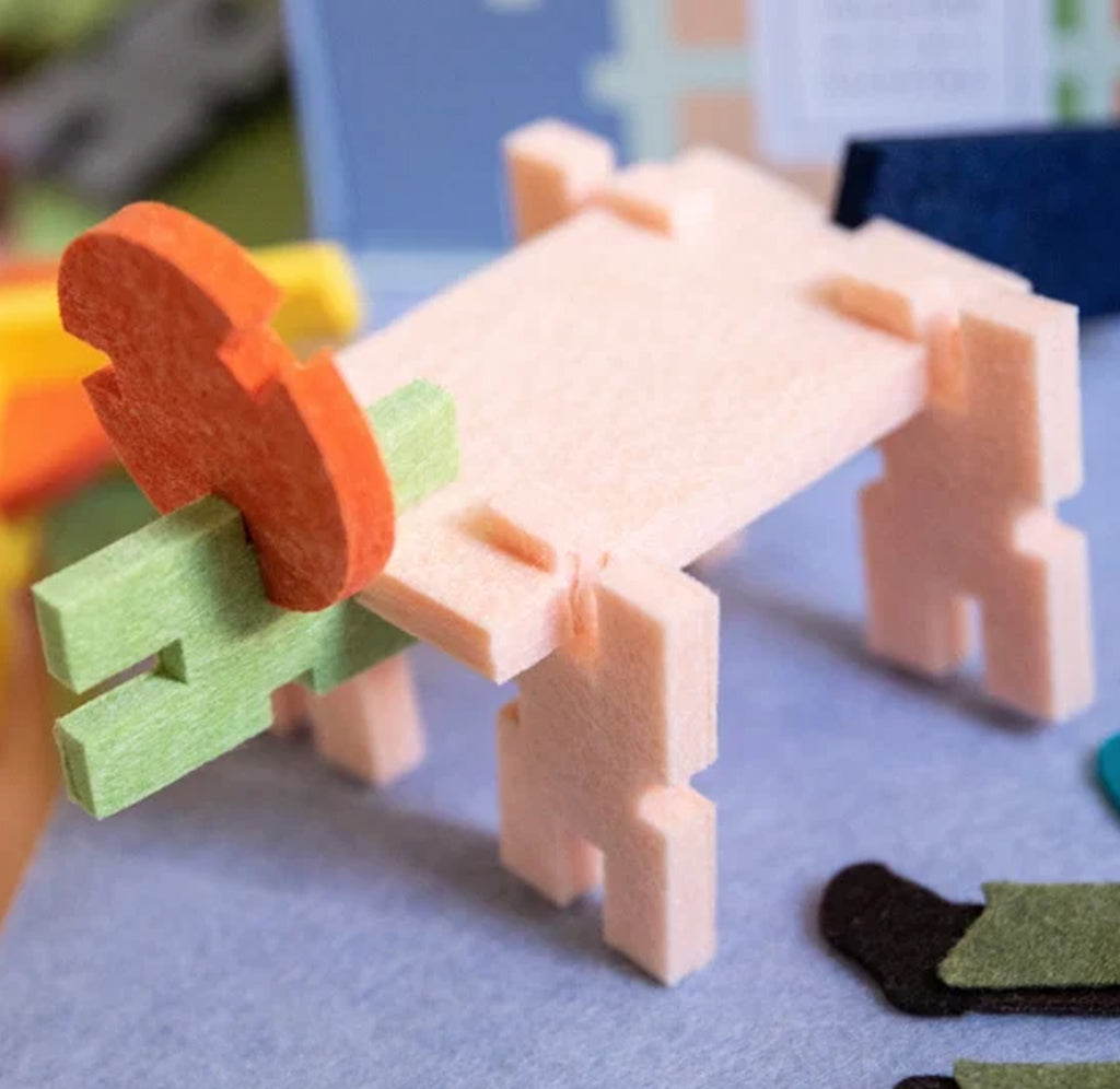 toy made from pale pink, light green, and orange felt pieces, with a hand holding one of the green felt pieces. The toy is set against a soft-focus blue and gray background.