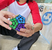 Person in a red-and-white shirt holds a colorful, scrambled dodecahedron puzzle cube, showing green, blue, and purple faces outdoors.