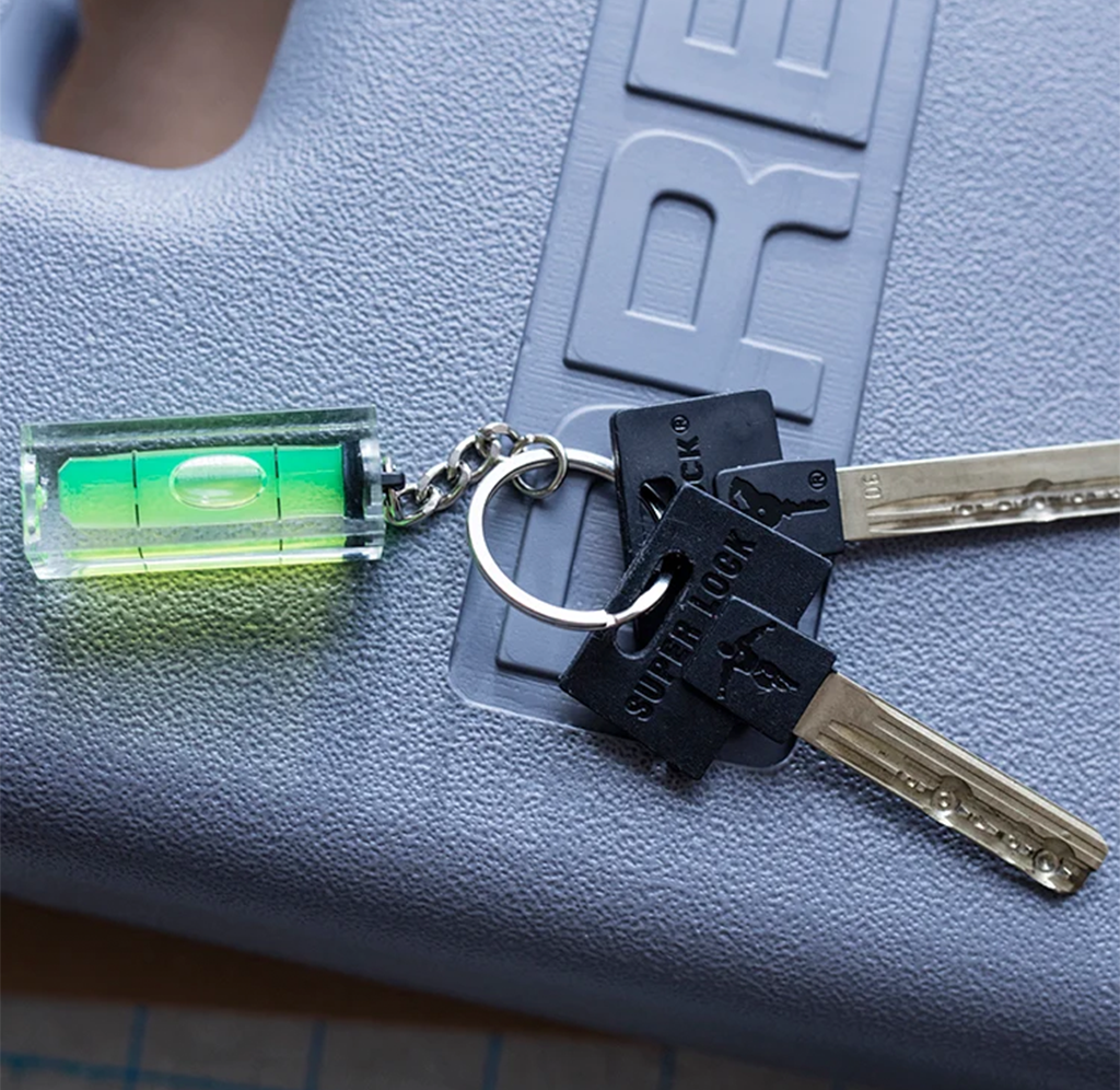 A small green bubble level on a key ring lies beside two metal keys on a textured gray surface, suggesting a precise, practical tool for leveling.