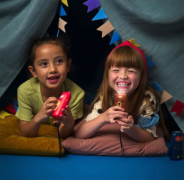 Two children smiling inside a cozy play tent, holding colorful Kidoki squeeze flashlights during indoor playtime.