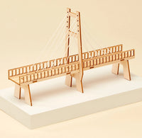 A wooden cable-stayed bridge model displayed on a white rectangular pedestal against a warm beige background.