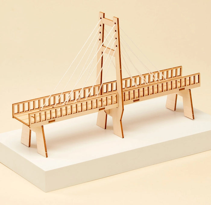 A wooden cable-stayed bridge model displayed on a white rectangular pedestal against a warm beige background.
