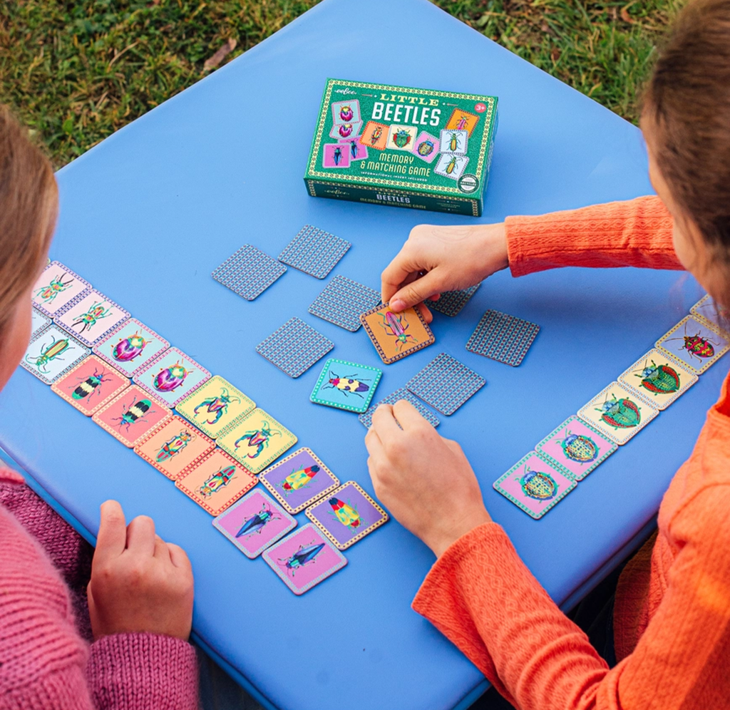 Two children playing a beetle-themed card game outdoors on a bright blue table. The box for the game is visible at the top, along with several colorful cards laid out on both sides and a few cards being placed in the center.