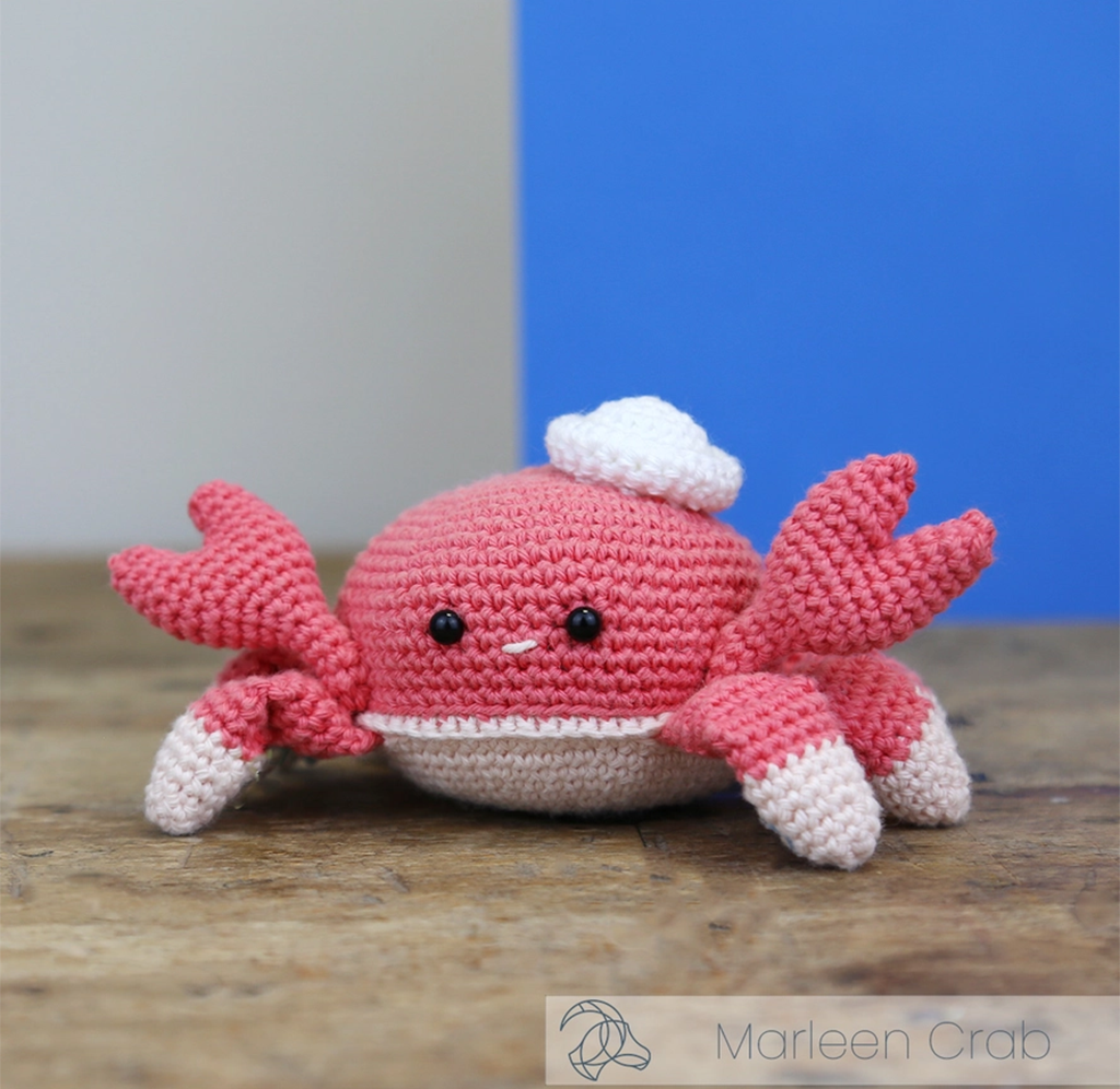 A cheerful pink crocheted crab plush with raised claws, white-tipped legs, bead eyes, and a tiny smile sits on a wooden surface against a blue-and-gray backdrop.