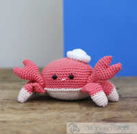 A cheerful pink crocheted crab plush with raised claws, white-tipped legs, bead eyes, and a tiny smile sits on a wooden surface against a blue-and-gray backdrop.