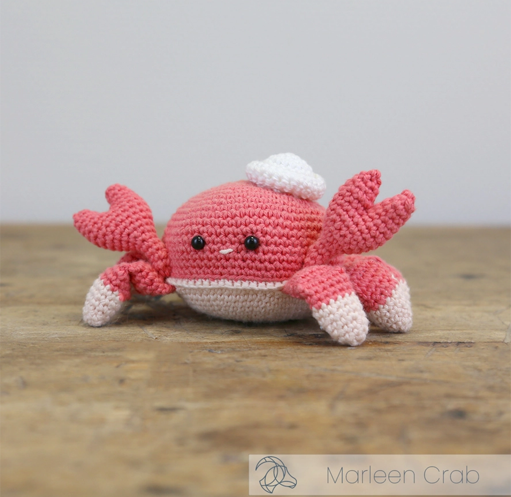 A cheerful pink crocheted crab with raised claws, black bead eyes, a white belly, and a tiny  hat sits on a wooden table.