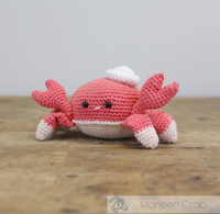 A cheerful pink crocheted crab with raised claws, black bead eyes, a white belly, and a tiny  hat sits on a wooden table.