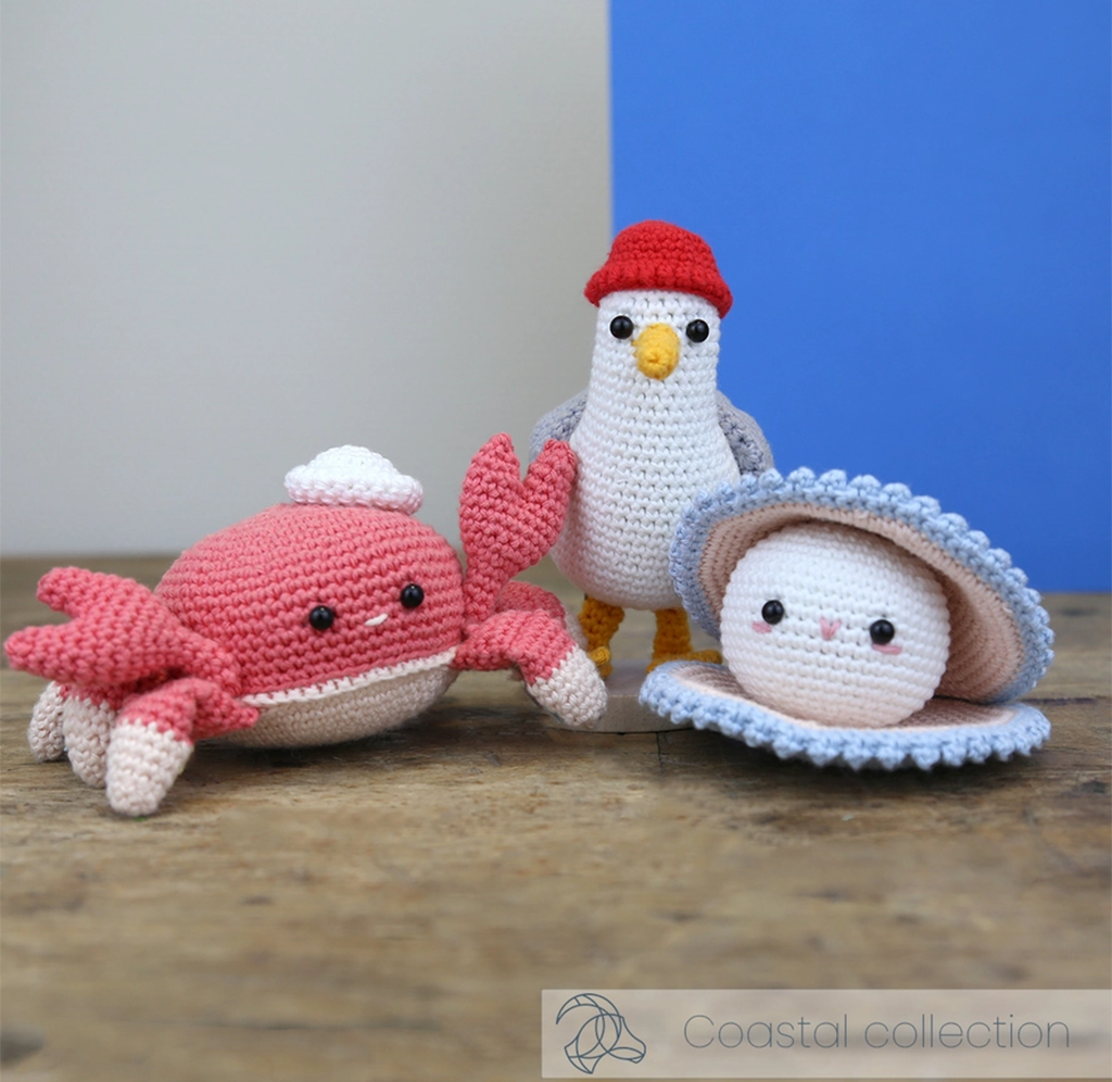 Crocheted coastal sea creatures—a red crab, white seabird in a red cap, and smiling shell—sit on a wooden surface against blue and gray walls, evoking a playful, handmade mood.