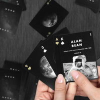 A close-up of a person's hands holding a fan of custom playing cards with a black and white moon theme. The top card is an Ace of Spades with 'ALAN BEAN' and 'APOLLO 13' text, and the card backs feature a detailed moon graphic.