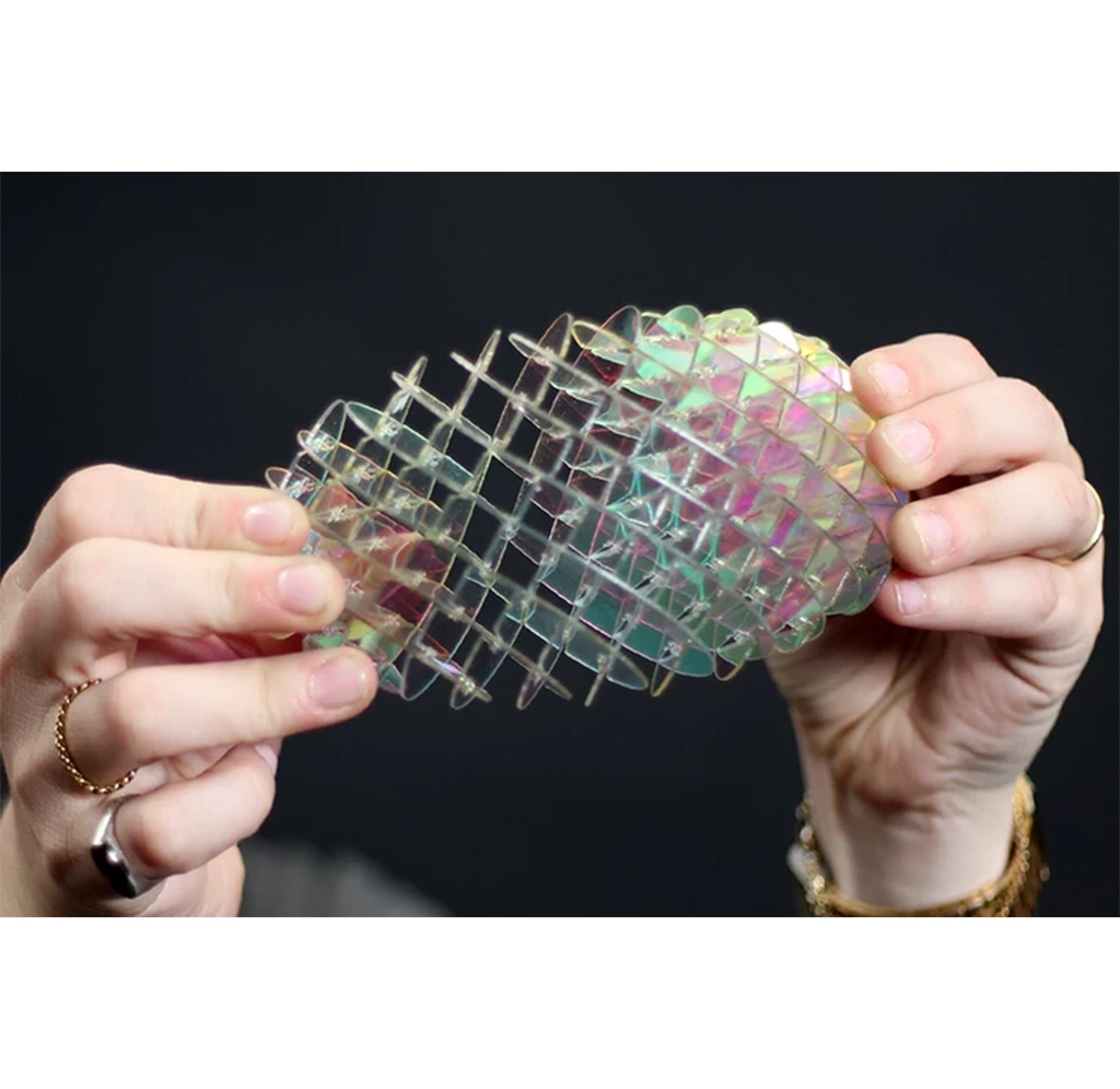 A close-up of a person's hands holding a flexible, translucent grid-like mesh object with iridescent pastel colors against a dark background.