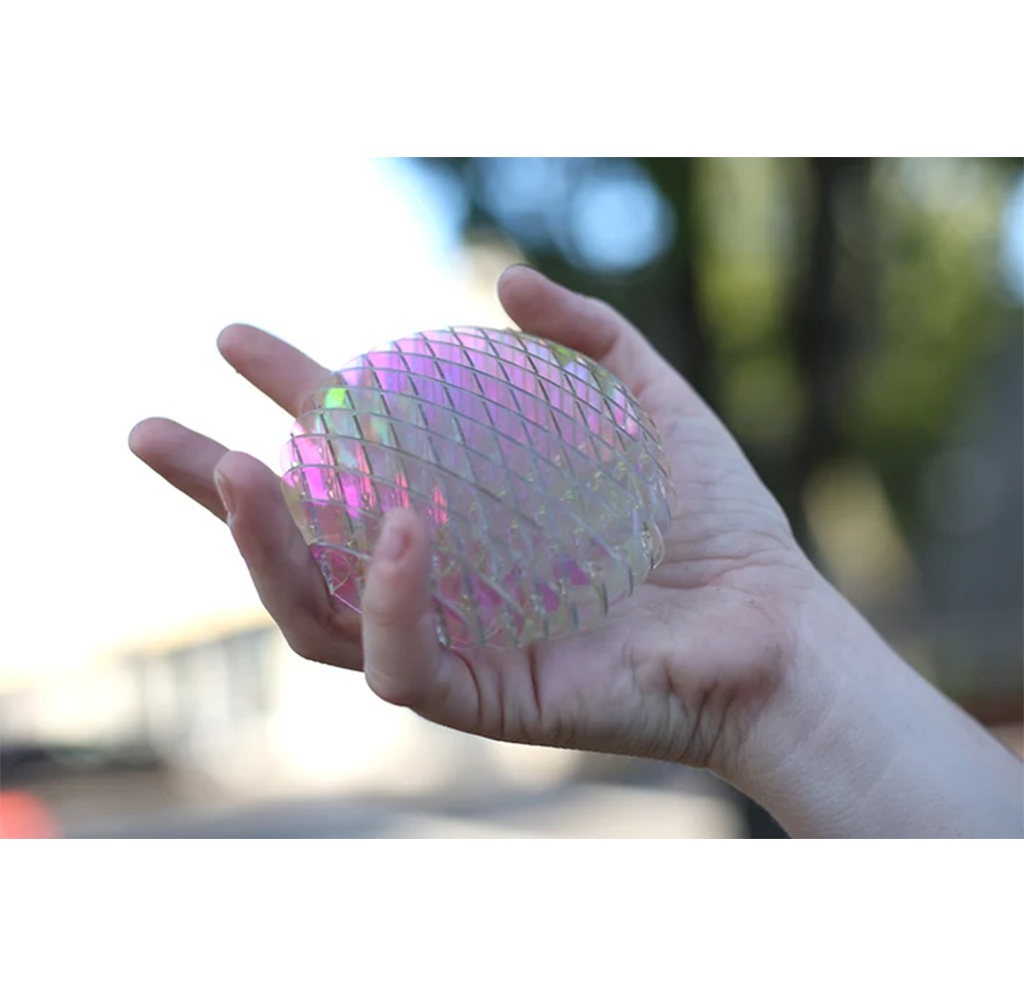 A hand holding a translucent, diamond-patterned fidget toy with iridescent pink and green reflective qualities, set against a blurred outdoor background.