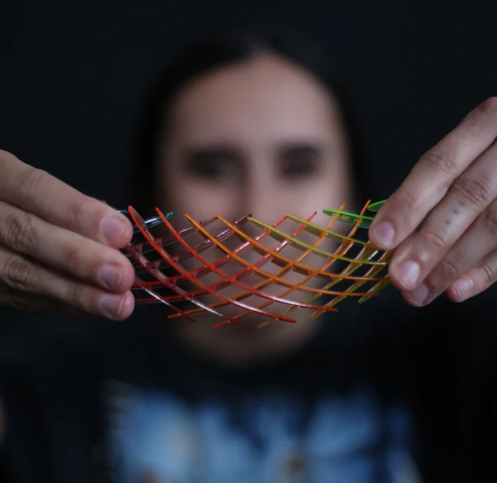 A close-up of a person's hands holding a flexible, crisscrossed mesh structure made of translucent orange, red, and green rods, with their face blurred in the background against a dark backdrop.