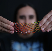 A close-up of a person's hands holding a flexible, crisscrossed mesh structure made of translucent orange, red, and green rods, with their face blurred in the background against a dark backdrop.