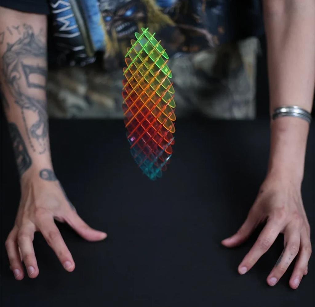 A person's tattooed arms resting on a black surface, with a translucent grid-like decorative object floating in the center. The object has a rainbow gradient transitioning from green and yellow at the top to orange and blue at the bottom.