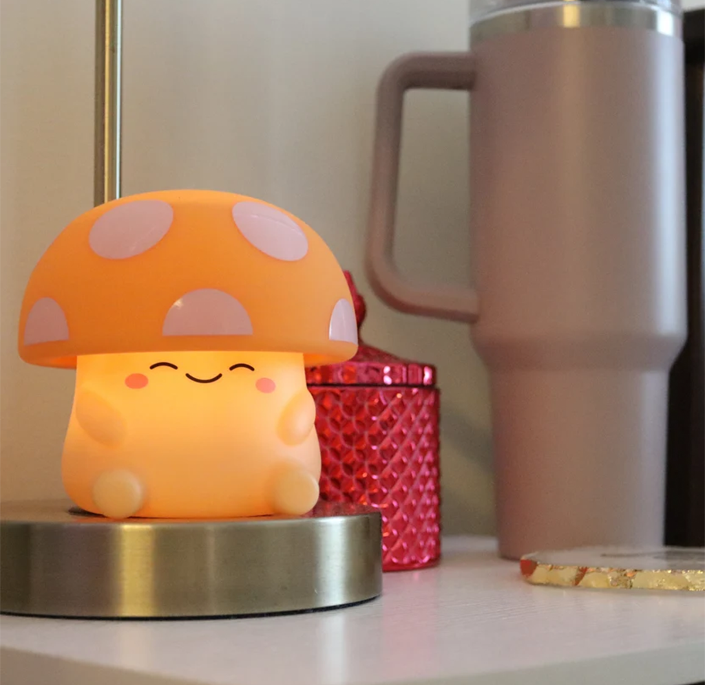 A cheerful orange mushroom-shaped lamp with a smiling face sits on a metal base beside a red textured container and tall travel mug.