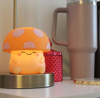 A cheerful orange mushroom-shaped lamp with a smiling face sits on a metal base beside a red textured container and tall travel mug.