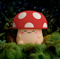 A cute, smiling mushroom figurine with a bright red cap and white spots sits among lush green moss in a shadowy forest, creating a whimsical, cozy mood.