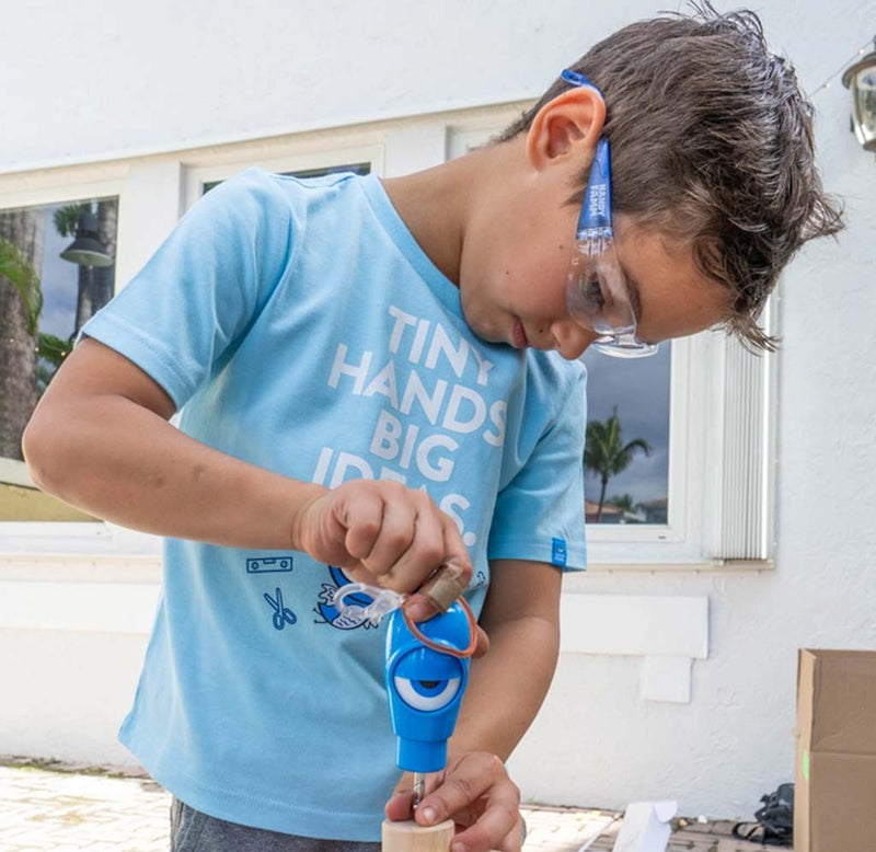 A young boy wearing safety glasses and a light blue graphic t-shirt is using a blue power drill to make a wooden object outdoors in front of a white building.