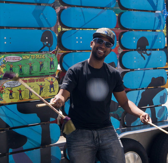 Male wearing a black short sleeve shirt with the eclipse optical illusion in white on the front. Exploratorium in white on the left shoulder.