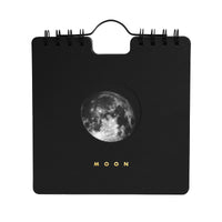 A black spiral-bound calendar with a circular moon graphic in the center and the word 'MOON' printed below in gold, shown on a plain white background