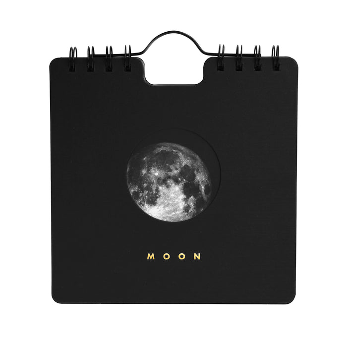 A black spiral-bound calendar with a circular moon graphic in the center and the word 'MOON' printed below in gold, shown on a plain white background
