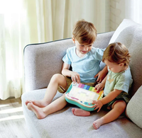 Two young siblings sit together on a gray sofa, smiling as they explore a colorful interactive piano in a bright, sunlit room; warm, affectionate mood.