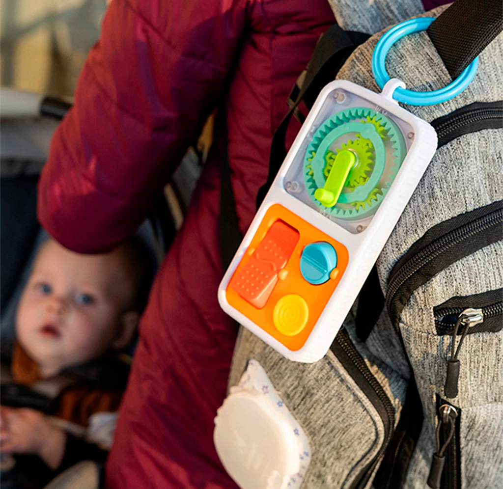 Close-up of a colorful sensory toy clipped to a gray backpack, with a baby in a stroller behind it; bright textures suggest playful, on-the-go entertainment.