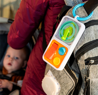 Close-up of a colorful sensory toy clipped to a gray backpack, with a baby in a stroller behind it; bright textures suggest playful, on-the-go entertainment.