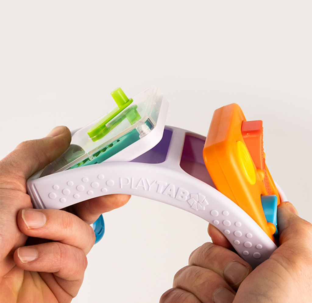 Hands stretch a flexible white PLAYTAB toy with bright green, orange, and blue components against a plain background, suggesting playful, hands-on fun.