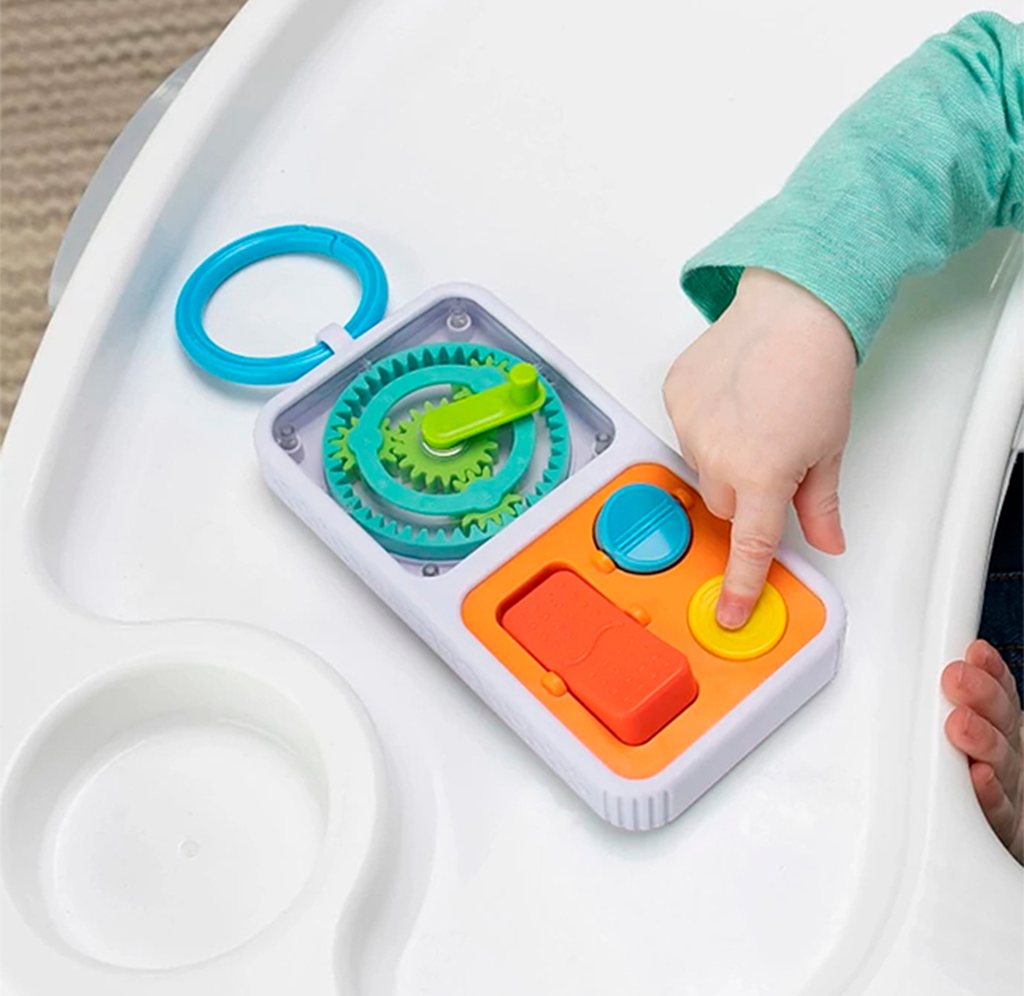 A child in a green sleeve presses a yellow button on a colorful gear-and-button activity toy attached to a white high-chair tray, exploring playfully.