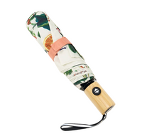 A compact, folded umbrella with a tropical fruit and leaf pattern on a white background, featuring a peach-colored strap and a light bamboo handle with a black logo.