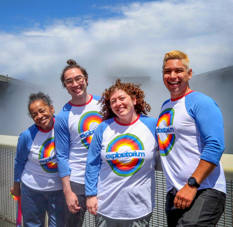 A group of four people is standing in front of the Exploratorium's Fog Bridge with "fog" spewing out behind them. They are all wearing the rainbow t-shirt, a white baseball tee with blue sleeves, and a circular rainbow design in the middle with Exploratorium in blue across the rainbow.