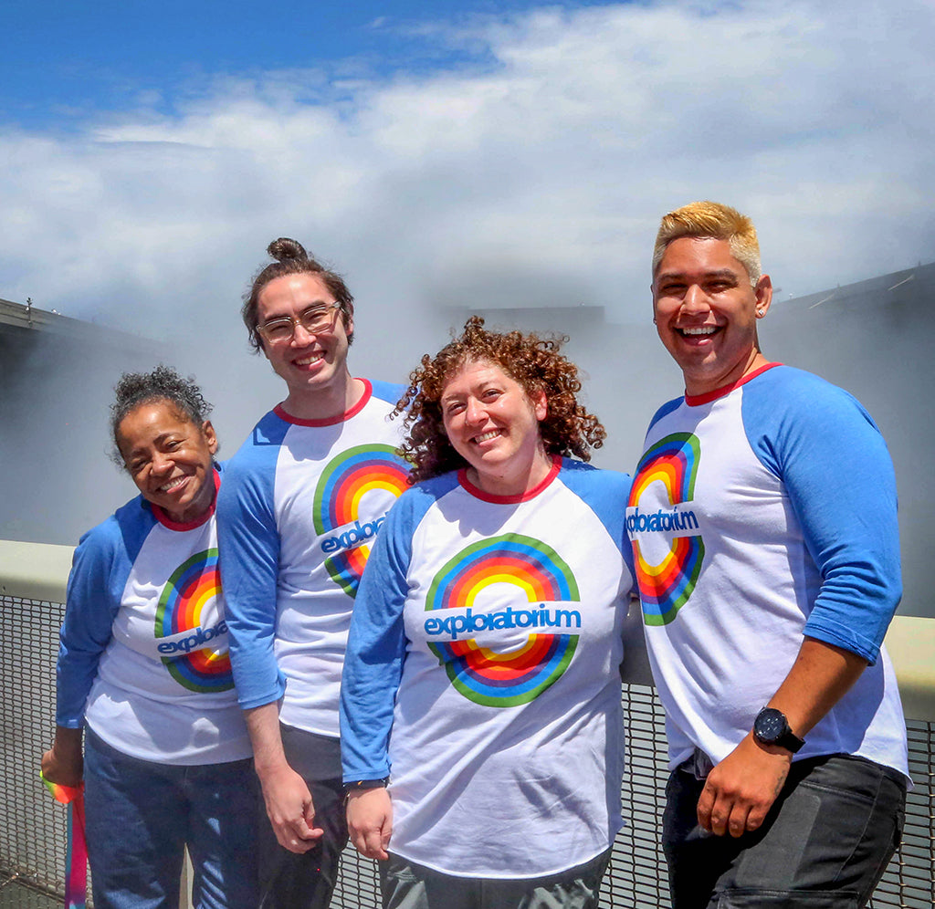 A group of four people is standing in front of the Exploratorium's Fog Bridge with "fog" spewing out behind them. They are all wearing the rainbow t-shirt, a white baseball tee with blue sleeves, and a circular rainbow design in the middle with Exploratorium in blue across the rainbow.