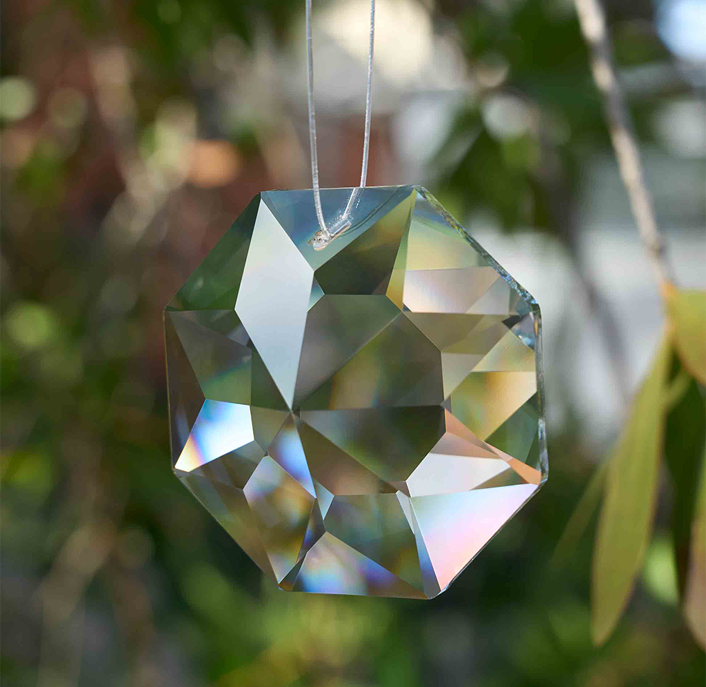 A faceted clear glass suncatcher ornament hanging from a thin cord, photographed outdoors against a blurred green and brown natural background.