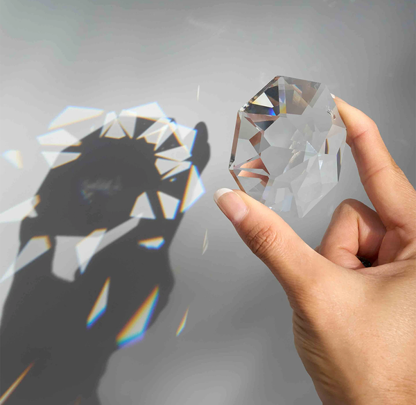 A hand holding a clear faceted crystal rainbow catcher that casts a dramatic shadow and rainbow light refraction on a gray background.
