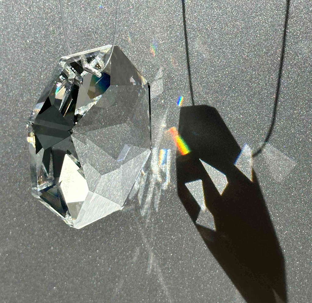 A clear faceted crystal prism casting sharp shadows and a rainbow spectrum on a textured gray surface.