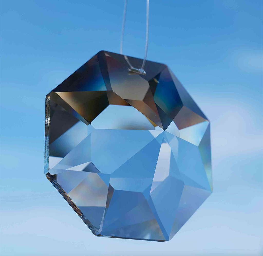 A faceted octagonal glass sun catcher with a hanging loop, shown against a soft blue sky gradient background.