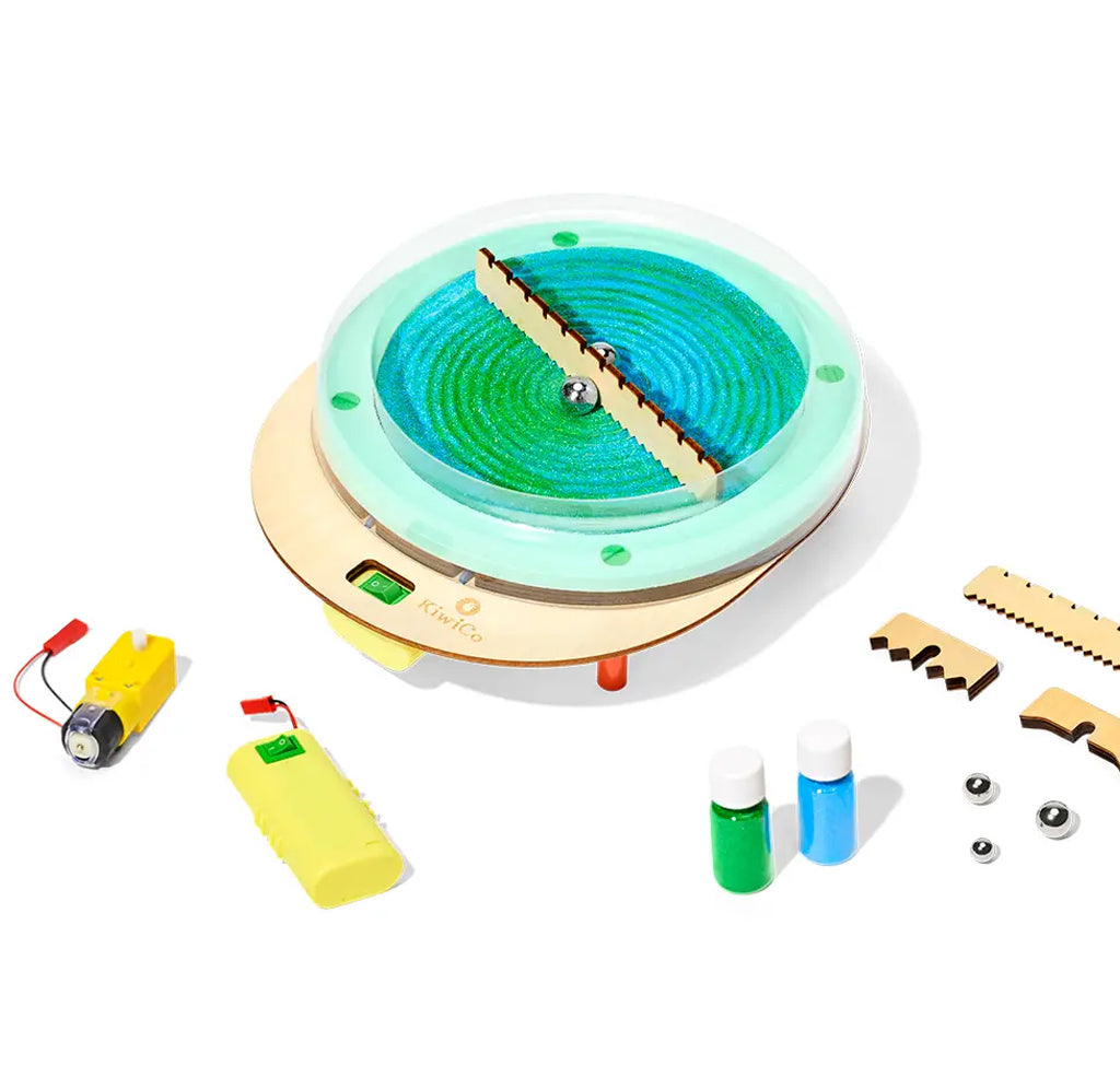 A children's science toy set on a white background, featuring a circular magnetic marbles in a base with a wooden slat, a yellow battery, a yellow motor, small bottles of green and blue sand, and wooden pieces.